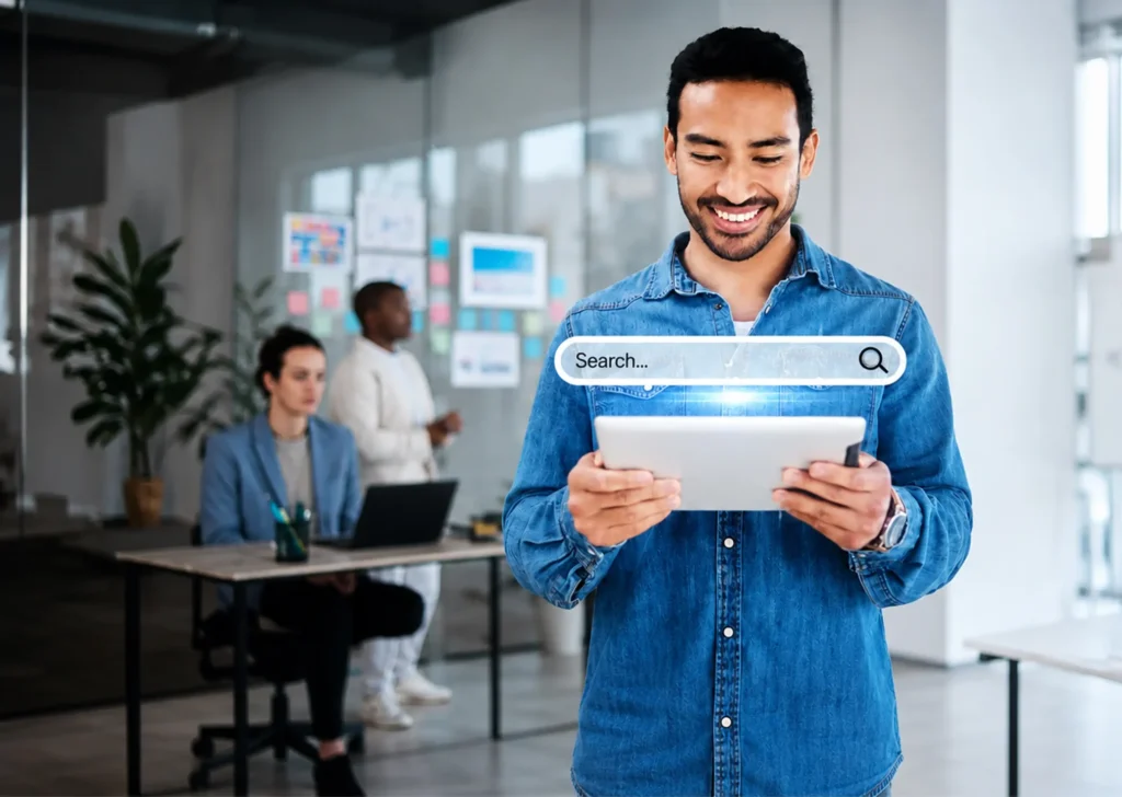 Person in a denim shirt holding a tablet with people in the background ad a virtual search bar above the tablet
